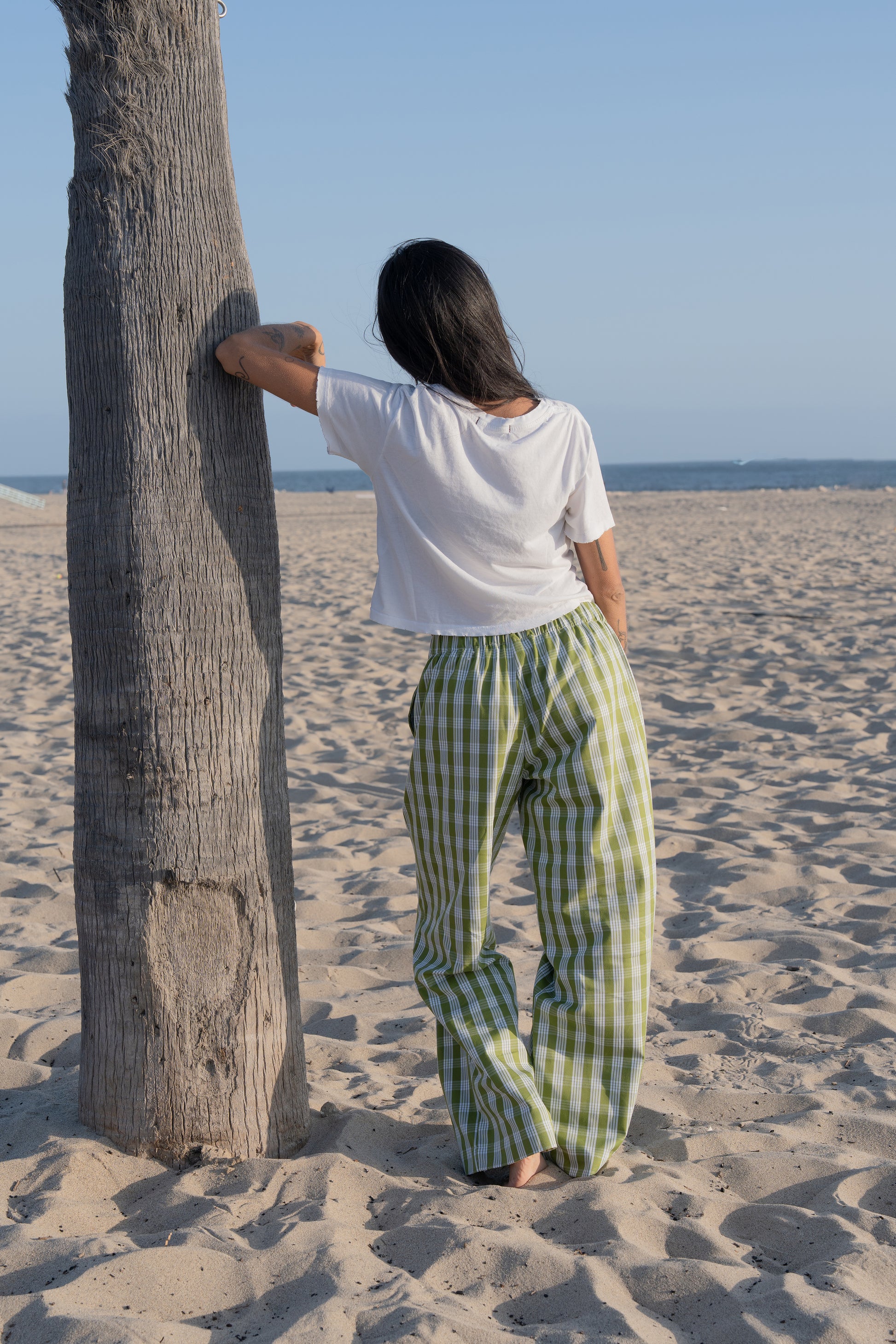 Woman in white top and palaka avo lounge pants by mostly sunny standing on a sandy beach with palm trees in the background.