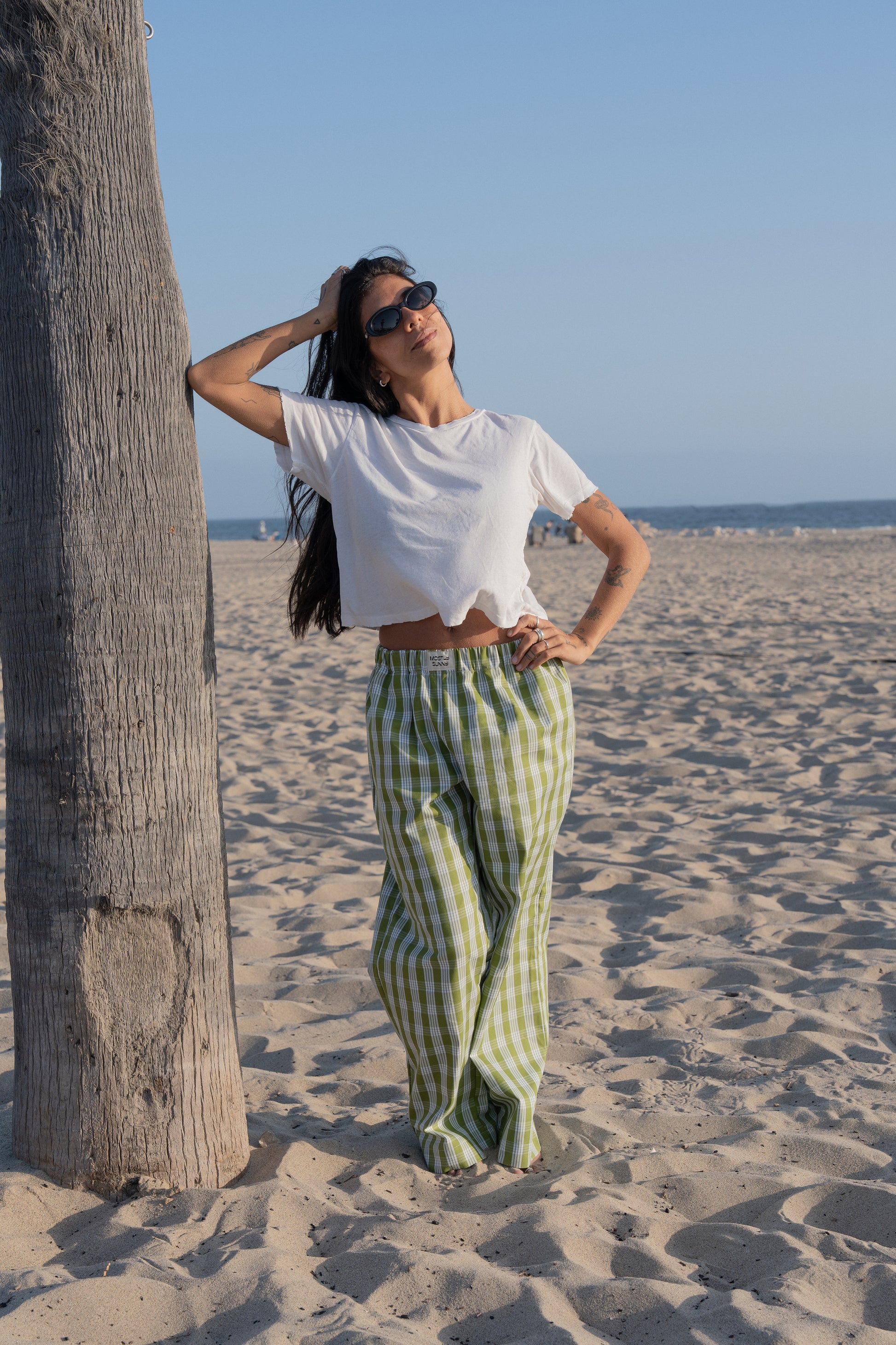 Woman in white top and palaka avo lounge pants by mostly sunny standing on a sandy beach with palm trees in the background.