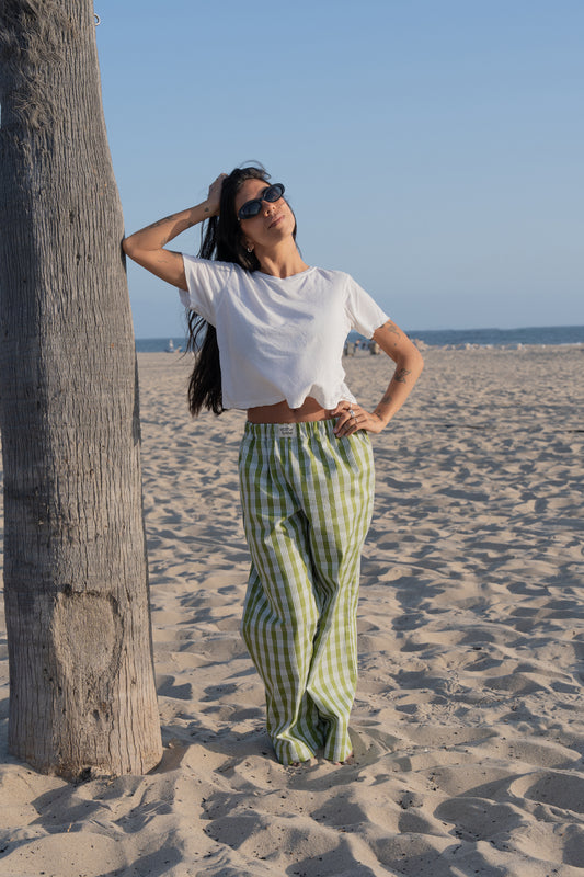 Woman in white top and palaka avo lounge pants by mostly sunny standing on a sandy beach with palm trees in the background.