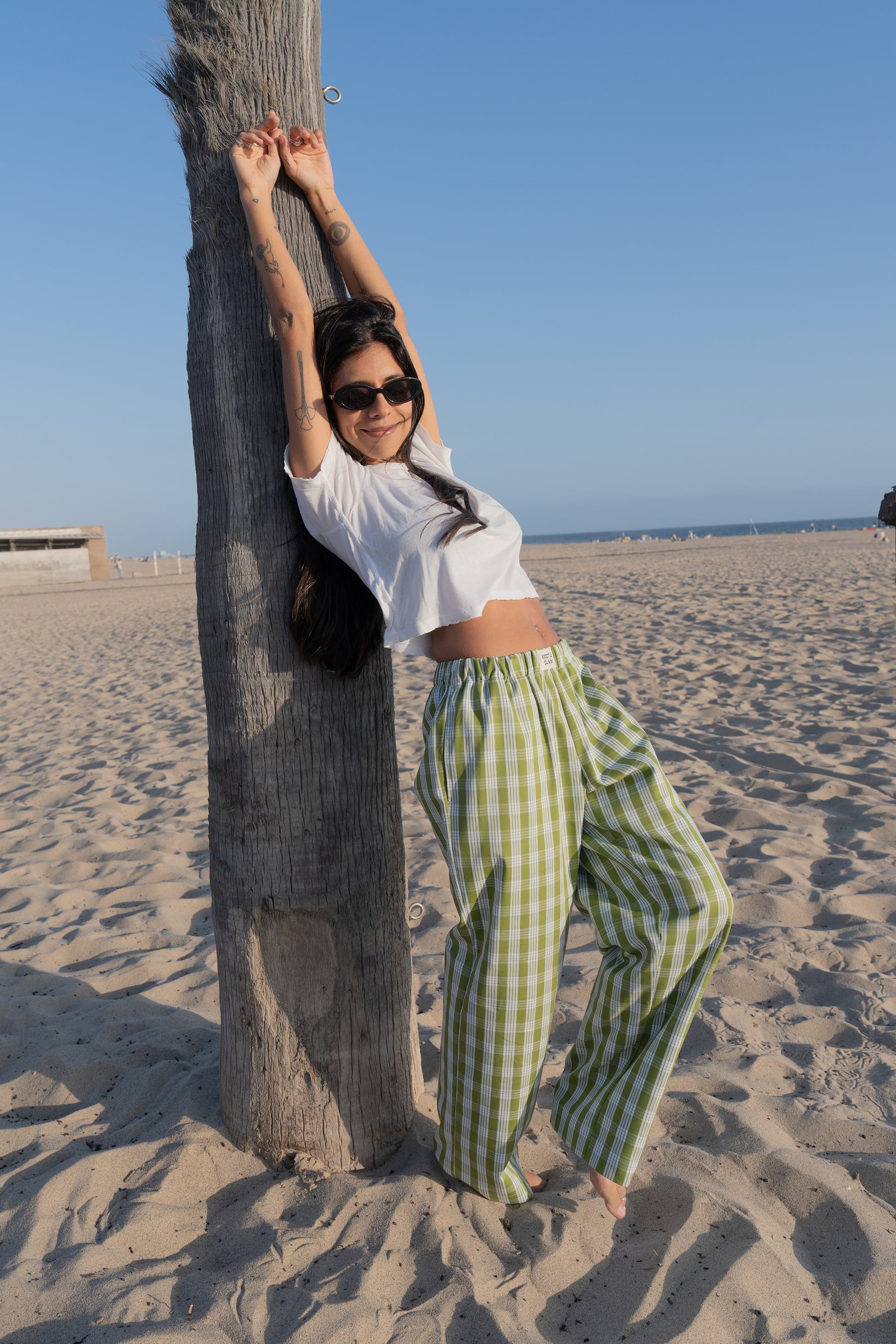 Woman in white top and palaka avo lounge pants by mostly sunny standing on a sandy beach with palm trees in the background.