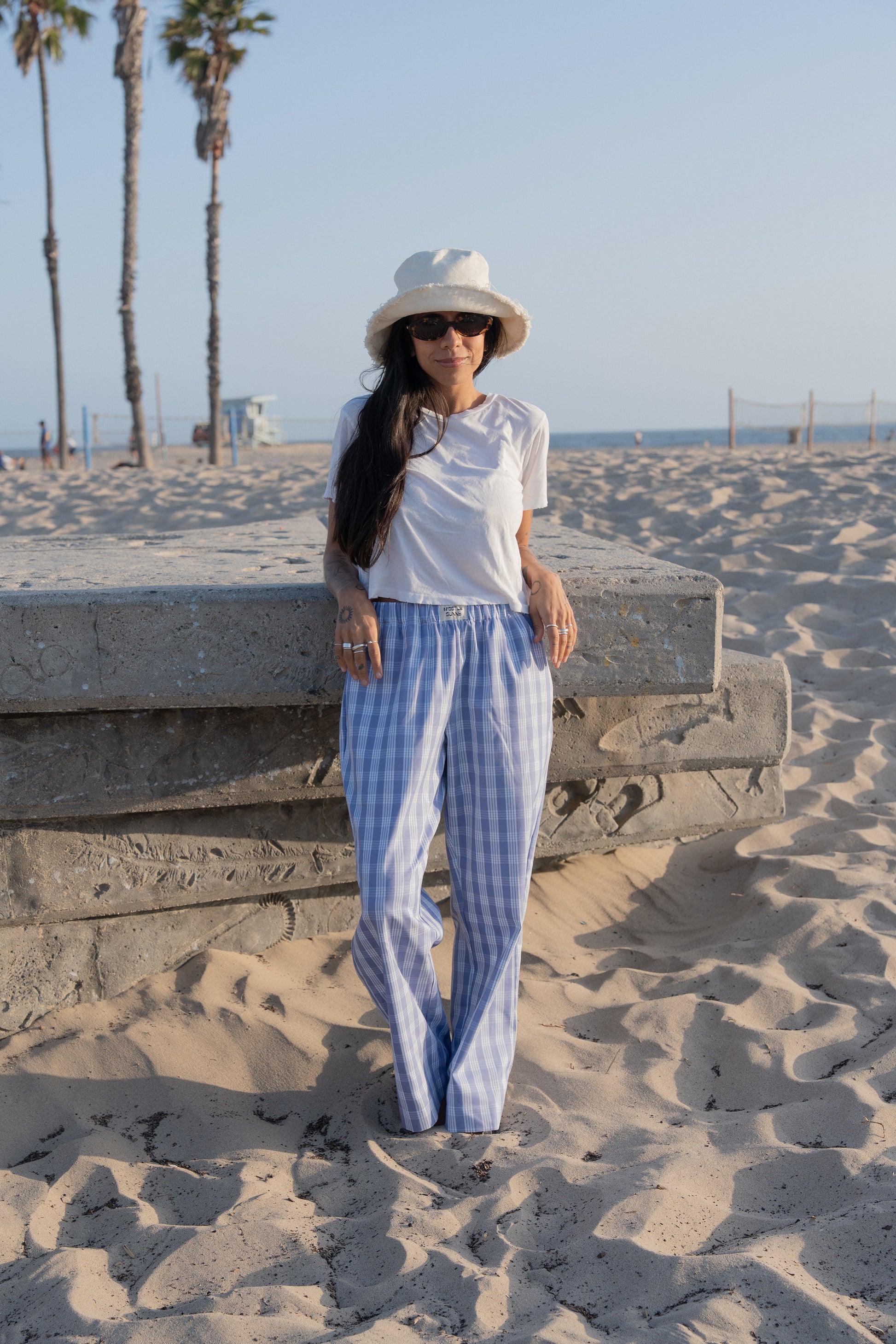 Woman in white top and palaka peri lounge pants by mostly sunny standing on a sandy beach with palm trees in the background.