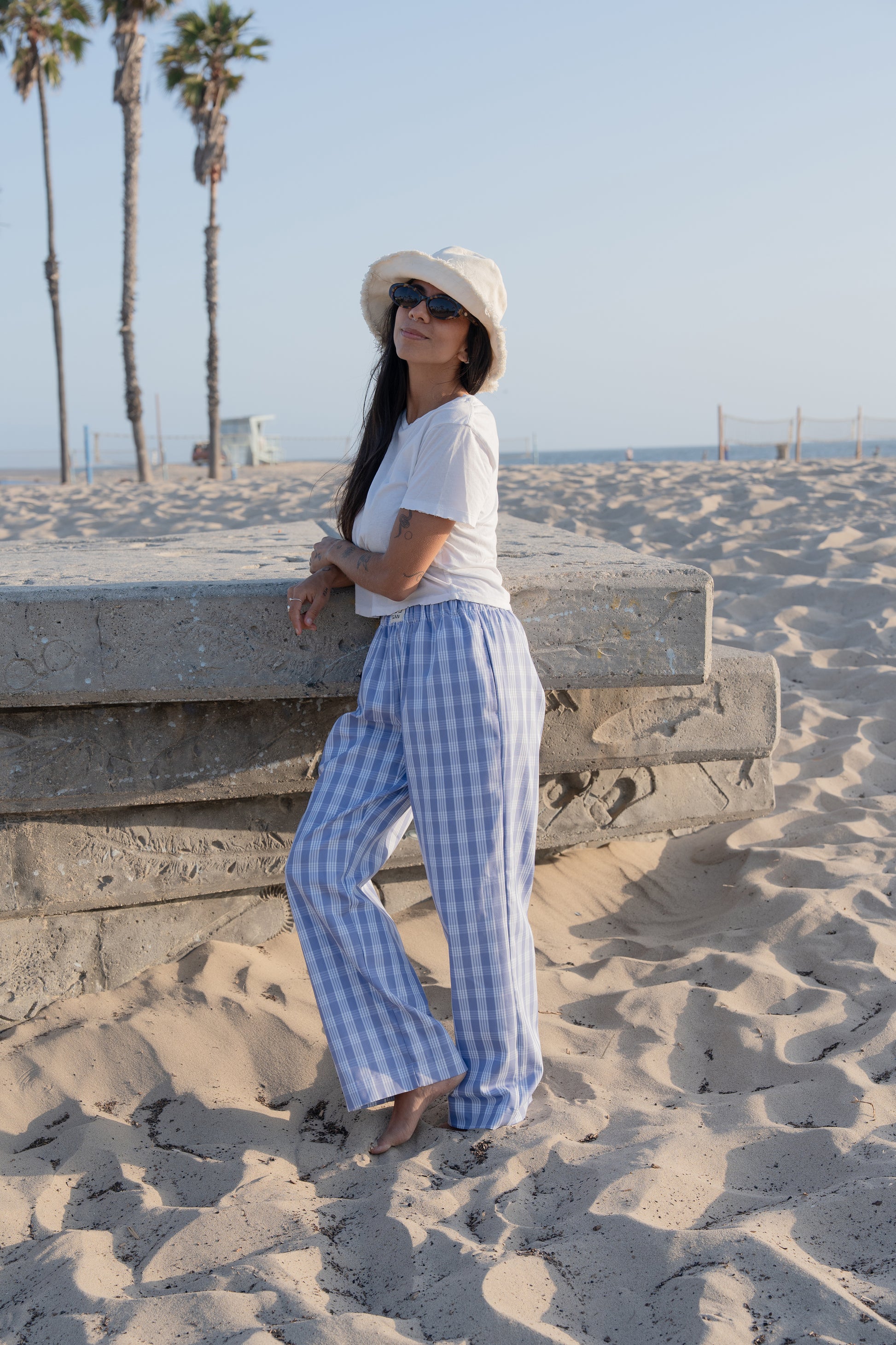 Woman in white top and palaka peri lounge pants by mostly sunny standing on a sandy beach with palm trees in the background.