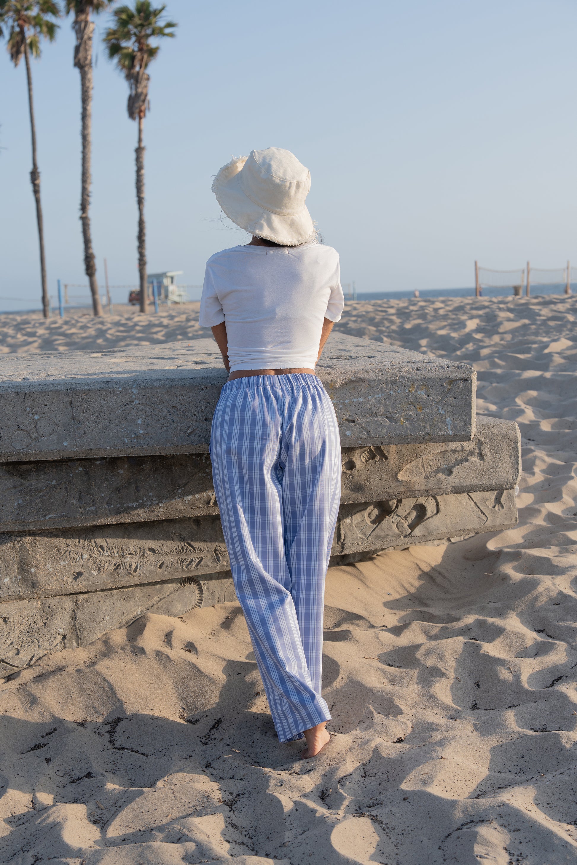 Woman in white top and palaka peri lounge pants by mostly sunny standing on a sandy beach with palm trees in the background.